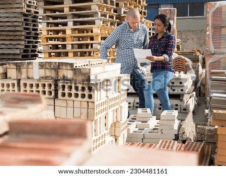 Confident woman manager discussing order list with man worker at warehouse of building materials