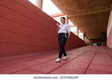 Confident woman in a pink shirt walking through modern building corridor while checking phone and holding a notebook - Powered by Shutterstock