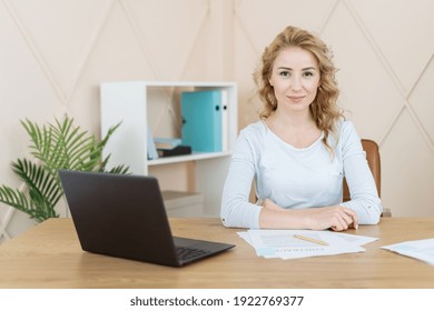 Confident Woman Sitting In Office Behind Desk With Laptop Computer And Documents, Looking At Camera. Smiling Businesswoman Or CEO Posing, Making Headshot Picture For Company Photoshoot