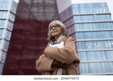 A confident woman stands in front of modern architecture wearing a tan coat in an urban setting during sunset - Powered by Shutterstock