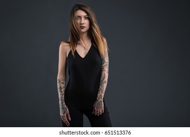 Confident Woman Studio Portrait With Long Black Dress And Tattoos Against Dark Background.