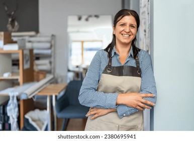 confident woman wearing a checkered shirt and beige apron stands in a workshop with folded arms, smiling towards the camera. - Powered by Shutterstock