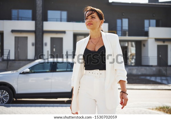 Confident woman in white formal wear outdoots near\
her white car.