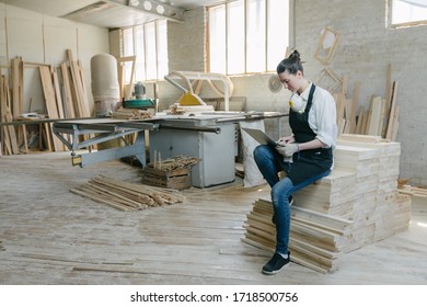 Confident Woman Working As Carpenter In Her Own Woodshop. She Using A Laptop And Writes Notes While Being In Her Workspace. Small Business Concept.