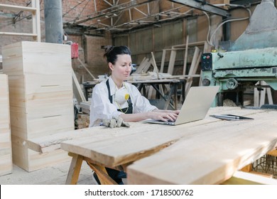 Confident Woman Working As Carpenter In Her Own Woodshop. She Using A Laptop And Writes Notes While Being In Her Workspace. Small Business Concept.