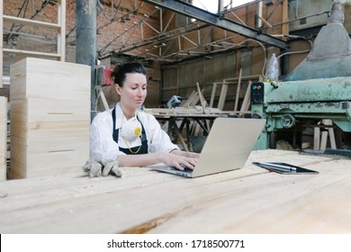 Confident Woman Working As Carpenter In Her Own Woodshop. She Using A Laptop And Writes Notes While Being In Her Workspace. Small Business Concept.