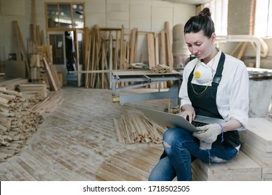 Confident Woman Working As Carpenter In Her Own Woodshop. She Using A Laptop And Writes Notes While Being In Her Workspace. Small Business Concept.