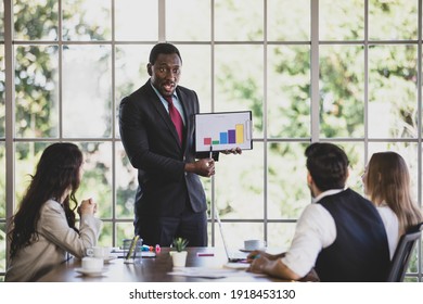 Confident Young Adult African Businessman Leader Presenting A Graph Document Of Their Company To Caucasian Colleagues In Meeting To Show The Financial Problem And Brainstorm Way To Solve It