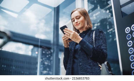 Confident Young Adult Female Riding Glass Elevator To Office In Modern Business Center. Successful Manager Using Smartphone, Write Message, Check Social Media And Emails In A Lift.