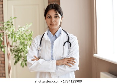 Confident Young Adult Indian Female Doctor Gp Wear White Coat Stethoscope Looking At Camera Standing With Arms Crossed In Medical Clinic Office. Health Care Professional Medic Physician Portrait.