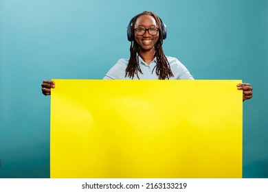 Confident young adult person with electronic earphones and empty advertisement placard. Happy positive woman holding yellow paper banner sign while having wireless modern headphones on blue background