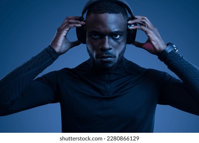 Confident young African American male athlete with dark hair and beard in activewear, adjusting headphones and looking at camera while listening to music during workout in blue studio - Powered by Shutterstock