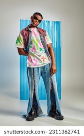Confident young african american man in colorful t-shirt, sunglasses and ripped jeans standing on grey with blue polycarbonate sheet at background, fashion shoot, sustainable lifestyle
