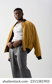 Confident young african american man in bomber jacket and pants posing and looking at camera isolated on grey, contemporary shoot featuring stylish attire, fashion statement