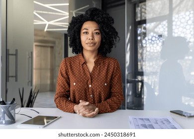 A confident young African American woman in a stylish polka dot shirt poses at her workspace with a tablet and blueprint ,in a well-lit modern office setting. - Powered by Shutterstock