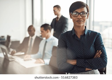 Confident young African businesswoman standing with folded arms smiling at the camera in a boardroom with male colleagues in the background - Powered by Shutterstock