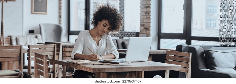 Confident young African woman writing something in note pad while working from cafe - Shutterstock ID 2270637409
