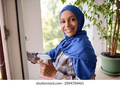 Confident young Arab Muslim cute woman with covered head in blue hijab, smiles looking at camera, washing windows, removing stains and wiping with rag during spring cleaning in house. Copy ad space