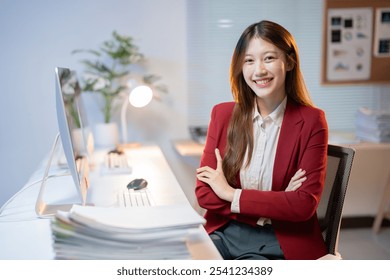 Confident young asian businesswoman smiling with arms crossed while sitting at her desk in a modern office, working late at night - Powered by Shutterstock