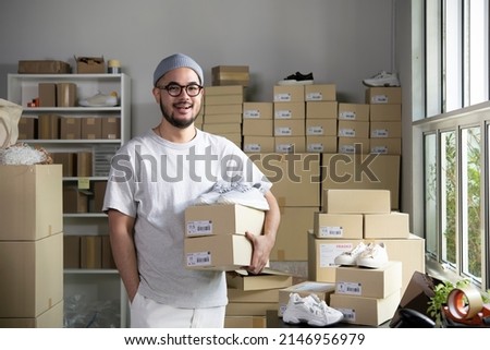 Confident young Asian man retail seller, entrepreneur, online store drop shipping small business owner looking at camera standing in delivery shipping warehouse with parcel boxes.