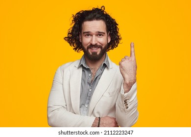 Confident young bearded male entrepreneur with wavy hair in formal clothes smiling at camera and showing index finger against yellow background