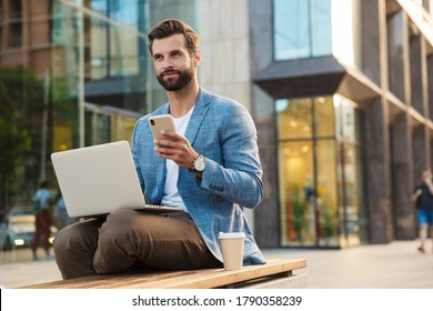 Confident Young Bearded Male Manager With Laptop And Smart Phone Sitting Outdoors