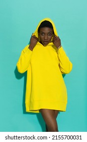 Confident young black woman in vibrant yellow oversized hoodie. Portrait against turquoise background. - Shutterstock ID 2155700031
