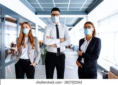 Confident young business man leader wearing a medical protective mask on his face, with a group of colleagues in the background, standing in an office building