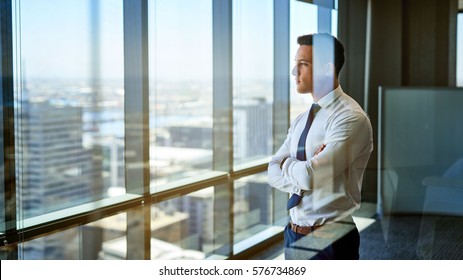 Confident young businessman deep in thought while looking through windows at the city from high up in an office building - Powered by Shutterstock