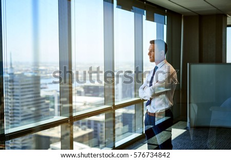 Confident young businessman deep in thought while looking through windows at the city from high up in an office building