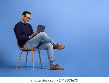 Confident young businessman in eyeglasses using laptop while sitting on chair over blue background