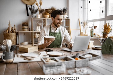 Confident young businessman running creative small business. Adult ceramic shop owner using laptop while working in store. Male entrepreneur managing online order of mug on website. - Powered by Shutterstock