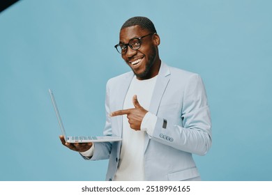 Confident young businessman smiling and pointing at laptop on blue background, dressed in light blue suit Modern professional concept for online business - Powered by Shutterstock