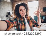 Confident young businesswoman with curly hair making an OK sign indoors in a modern office setting