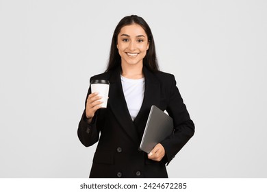 Confident young businesswoman smiling at camera, holding takeaway coffee cup in one hand and sleek laptop in the other, ready for work, grey backdrop - Powered by Shutterstock