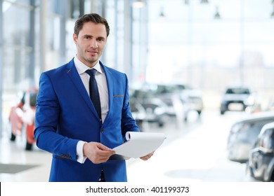 Confident Young Car Dealer Standing In Showroom.