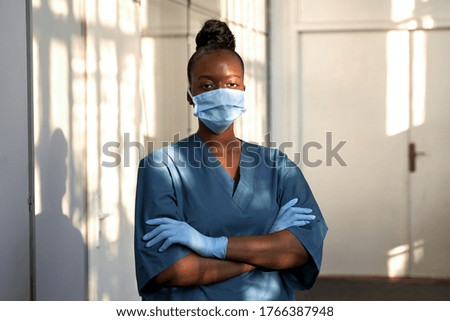 Confident young female african scrub nurse wear blue uniform, face mask, gloves standing arms crossed in hospital hallway. Black millennial woman doctor, surgeon, medic staff professional portrait.