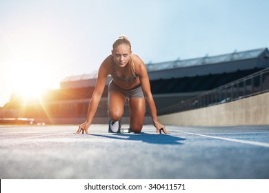 Confident young female athlete in starting position ready to start a sprint. Woman sprinter ready for a run on racetrack with sun flare. - Powered by Shutterstock