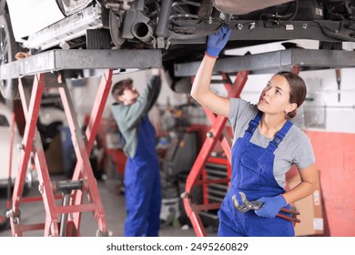 Confident young female auto mechanic in blue overalls standing under car on lift in service workshop, holding adjustable wrench and examining vehicle undercarriage - Powered by Shutterstock