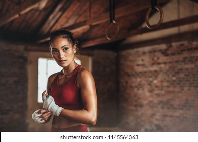 Confident young female boxer wearing strap on wrist. Boxing practice at gym. Woman in sports clothing preparing for boxing fight or workout. - Powered by Shutterstock