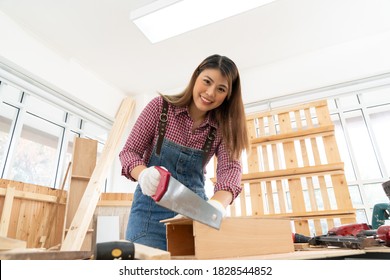 Confident Young Female Carpenter Cutting Wood With A Handsaw In The Carpentry Shop. Asian Handywoman Apprentice Sawing Wood In The Workshop. DIY Woodworking Crafts And Hobbies Concepts.
