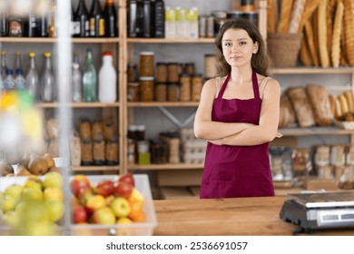 Confident young female grocery store clerk in burgundy apron smiling warmly, standing with arms crossed, surrounded by stocked shelves with freshly baked bread, fruits, and canned goods in cozy shop - Powered by Shutterstock