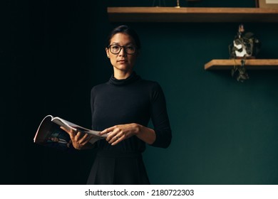 Confident young female holding magazine and looking at camera. Businesswoman standing with book in office. - Powered by Shutterstock