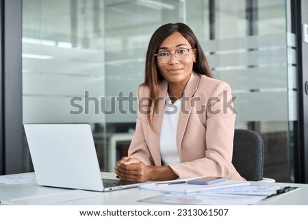 Confident young female lawyer, professional African American business woman company manager executive wearing suit glasses working on laptop in office sitting at desk looking at camera, portrait.
