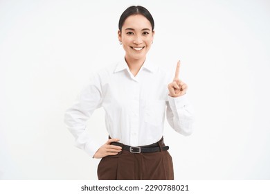 Confident young female manager, asian woman shows number one finger, explains smth, stands over white background.