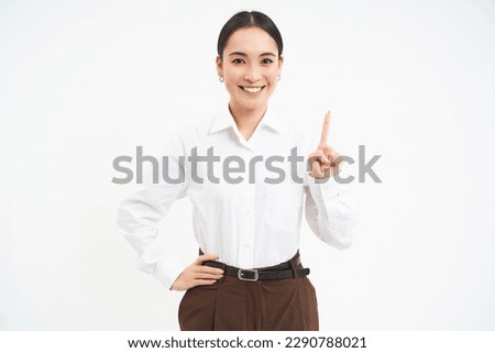 Confident young female manager, asian woman shows number one finger, explains smth, stands over white background.