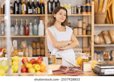 Confident young female seller in white uniform smiling welcomingly, standing with arms crossed at counter, surrounded by freshly baked goods, fruits, and various goodies in cozy grocery store - Powered by Shutterstock