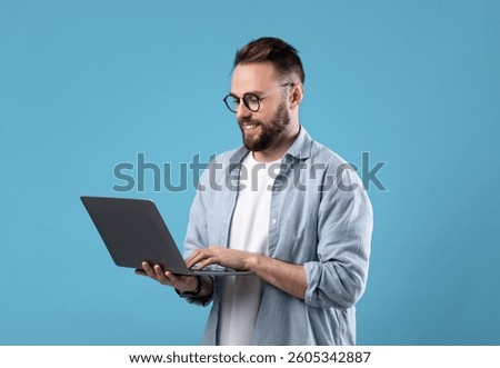 Confident young guy in glasses using laptop pc for online work or communication on blue studio background. Successful millennial guy participating in webinar, having remote meeting