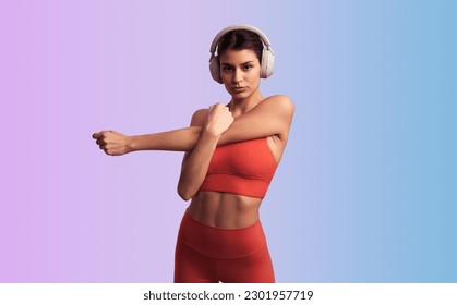 Confident young Hispanic female athlete with in trendy sportswear listening to music in wireless headphones, and looking at camera while stretching triceps against gradient background - Powered by Shutterstock