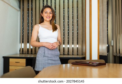 Confident Young Hispanic Female Entrepreneur Standing In Meeting Room Near Table With Folder Of Documents. Invitation To Negotiate Concept.